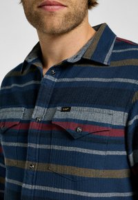 Camicia in flanella a quadri blu con trama a spina di pesce. Presenta strisce a contrasto in grigio, rosso e giallo. Due tasche sul petto con chiusura a bottone.