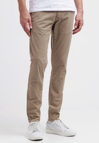 Beige slim fit chinos i bomull med slät struktur, fem fickor och bälteshällor, kombinerade med vita sneakers.