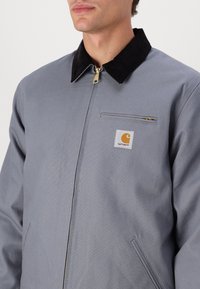 Veste grise en tissu durable, dotée d'un col en velours côtelé noir, d'une fermeture éclair à l'avant et d'un patch logo sur la poitrine gauche.