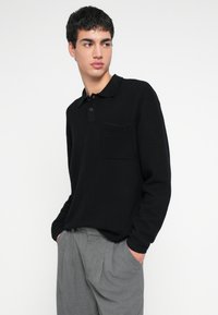 Μαύρο πόλο με μακριά μανίκια και υφή, με γιακά και τσέπη στο στήθος, συνδυασμένο με ανοιχτό γκρι πιετετέ παντελόνι.