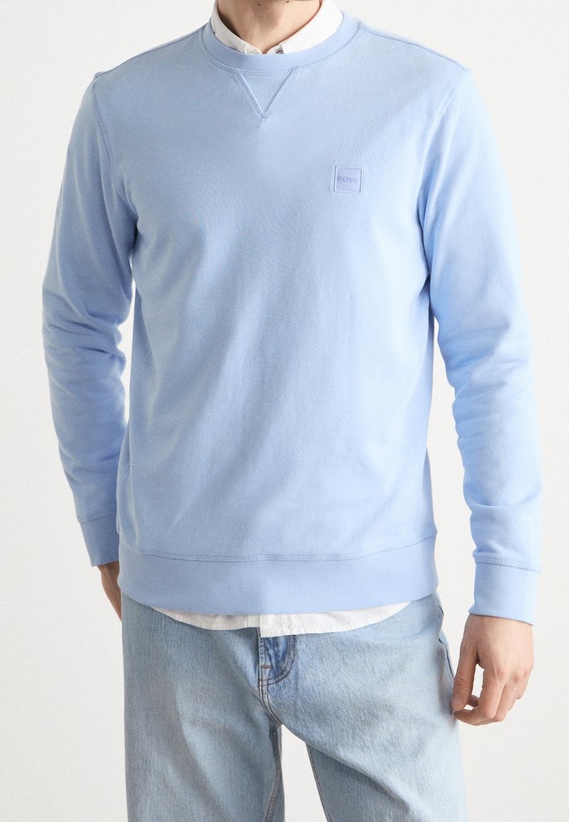 Homme portant un pull bleu clair à col rond par-dessus une chemise blanche et un jean bleu clair, debout devant un fond uni.