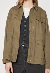 Kobieta w oliwkowej kurtce na czarnym, zapinanym kamizelce oraz w czarnych dżinsach z wysokim stanem, z białą bluzką pod spodem.