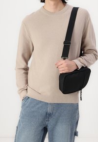 Persoon in een beige gebreide trui en blauwe spijkerbroek die een zwarte schoudertas met een dikke band vasthoudt tegen een witte achtergrond.