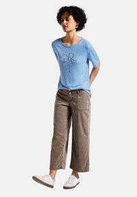 Camiseta de mangas cortas azul claro con un estampado gráfico, combinada con pantalones cortos marrones. Zapatillas blancas informales completan el conjunto.