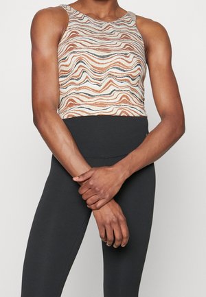 Person iført ærmeløs crop top med bølget mønster i beige toner og høj-taljede sorte leggings, stående med hænderne foldet.