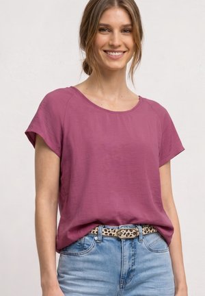 Smilende kvinde med lyst brunt hår i lav knold iført en kortærmet lilla top, blå jeans og et bælte med leopardprint mod en ensfarvet baggrund.