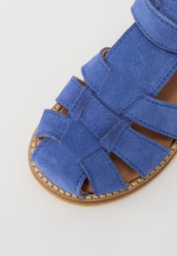 Sandal de camurça azul com tiras cruzadas e design de ponta aberta. Sola castanho-clara com costura visível à volta da borda.