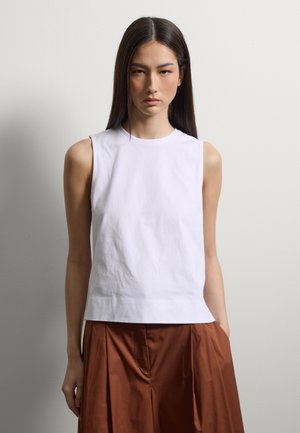 Junge Frau mit glatten dunklen Haaren, die ein ärmelloses weißes Top und lockere braune Hosen vor einem einfarbigen hellen Hintergrund trägt.