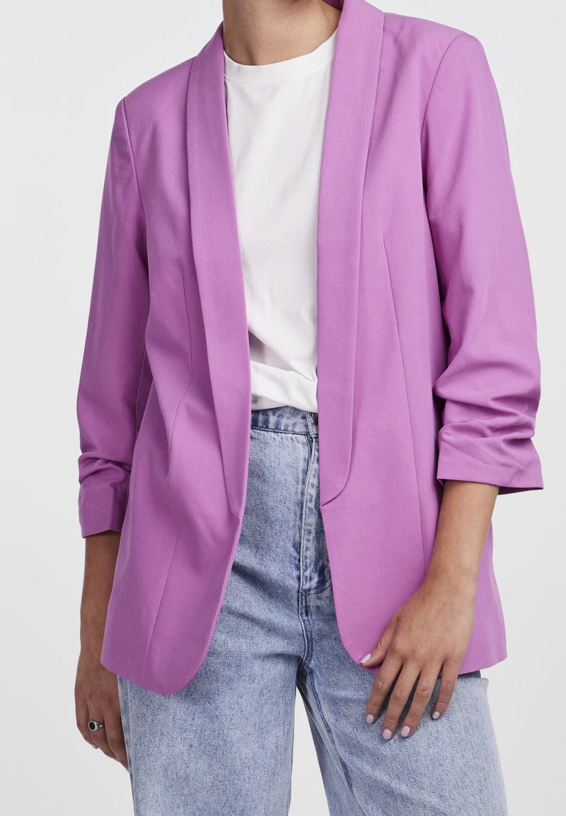 Personne portant un blazer lavande à manches retroussées par-dessus une chemise blanche et un jean bleu clair, debout devant un fond uni.