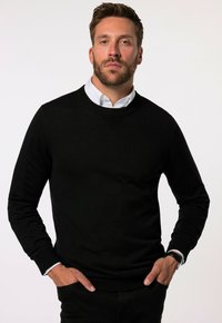 Maglione nero a maglia con collo rotondo, caratterizzato da un design testurizzato sul colletto, indossato sopra una camicia bianca con colletto. Mani inserite nelle tasche.