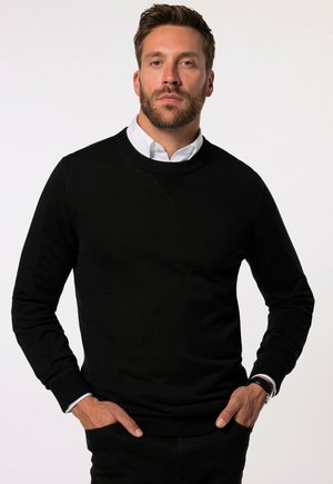 Maglione nero a maglia con collo rotondo, caratterizzato da un design testurizzato sul colletto, indossato sopra una camicia bianca con colletto. Mani inserite nelle tasche.