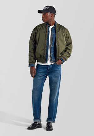 Joven con una chaqueta bomber verde oscuro sobre una chaqueta vaquera azul y camiseta blanca, vaqueros azules, zapatos negros y una gorra de béisbol negra.