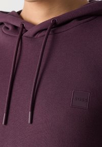 Gros plan d'une personne portant un sweat à capuche violet foncé avec des cordons et un petit patch carré portant l'inscription "BOSS" sur la poitrine.