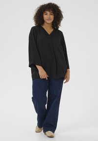 Black V-neck blouse with loose fit, long sleeves, and soft texture, paired with wide-leg blue jeans and beige shoes. Plain white background.
