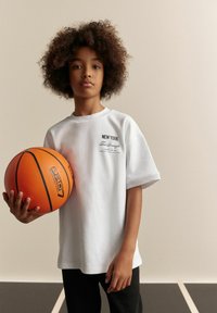 Hvid kortærmet T-shirt med teksten "NEW YORK", parret med sorte bukser. Holder en orange basketball med sorte linjer og logo.
