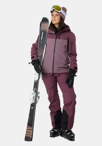 Skioutfit i lilla og sort, med en vandtæt jakke med lynlåslommer, matchende bukser, skibriller og ski med bindinger.