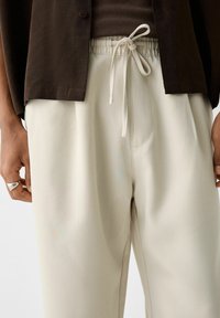 Béžové volné kalhoty s hladkou texturou, elastickým páskem a šňůrkou, doplněné tmavě hnědou košilí na knoflíky.