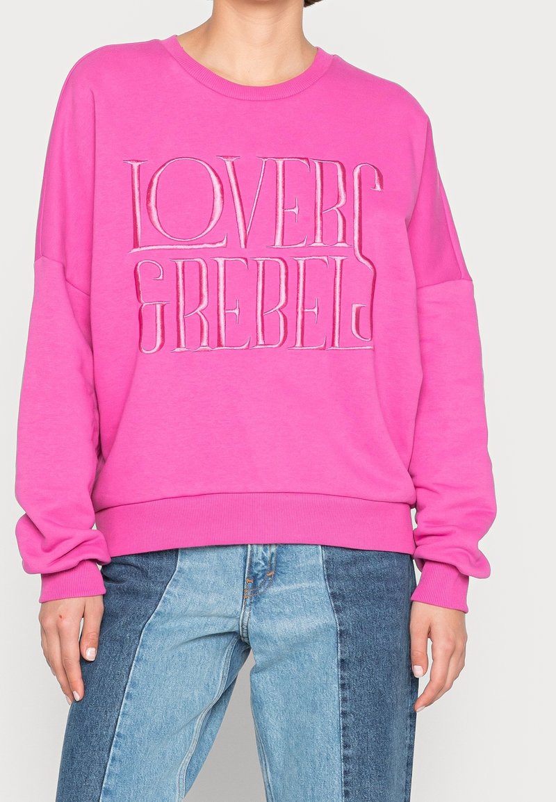 Personne portant un sweat-shirt rose vif avec le texte "LOVERS REBELS" et un jean bicolore bleu, debout devant un fond clair.