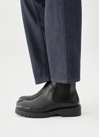 Fekete bőrbokacipő sima felülettel, rugalmas oldalpanelekkel és masszív talppal, sötét farmernadrággal párosítva.