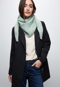 Lysegrøn uldsjaal med et struktureret mønster hængende om halsen, parret med en grå top og en mørk frakke. Jeans synlige ved taljen.