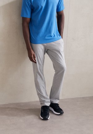 Mann trägt ein blaues Kurzarmhemd, hellgraue Jogginghose mit Logo und schwarze Sportschuhe und steht an einer beigen Wand.