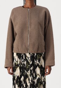 Žena v hnědém fleecovém zipovém saku s texturou přes černými, bílými a olivovými abstraktními vzory s knoflíky.