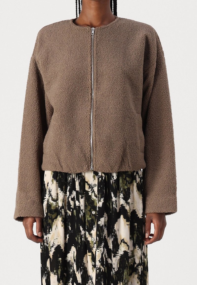 Žena v hnědém fleecovém zipovém saku s texturou přes černými, bílými a olivovými abstraktními vzory s knoflíky.