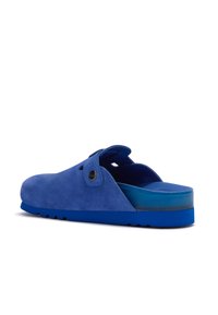 Zoccoli blu in suede con cinturino regolabile e suola testurizzata, angolati per mostrare la vista laterale e posteriore su uno sfondo bianco.