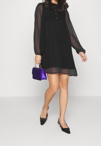 Black dress with sheer long sleeves and decorative embroidery, paired with a small purple handbag and black pointy-toed shoes.
