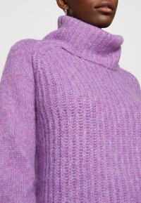 Gros plan sur une personne portant un pull à col roulé lavande doux à côtes, avec une texture duveteuse, et une petite boucle d'oreille visible.