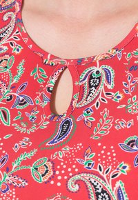 Červený top z látky s kulatým výstřihem a malým klíčovým otvorem, zdobený barevnými paisley a květinovými vzory.