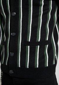 Cárdigan negro con rayas verticales verdes y blancas, tejido texturizado y cinco botones negros. Incluye un bolsillo en la parte frontal.