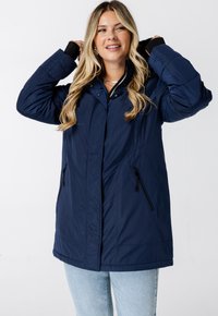 Navyblauwe geïsoleerde jas met een ritssluiting aan de voorkant, twee zijzakken met rits en een hoge kraag. Voorzien van lange mouwen met zwarte accenten.