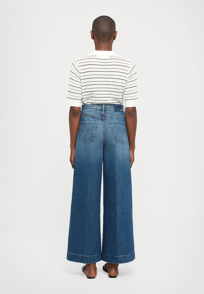 Personne debout de dos, portant un haut à manches courtes blanc à motifs, un jean bleu large et des chaussures plates noires.