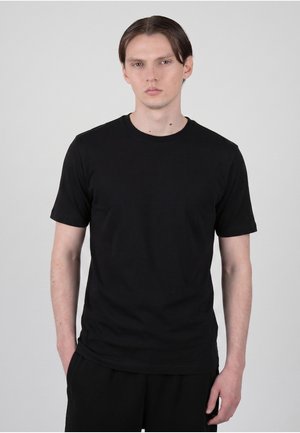 Ung mand med glat brunt hår iført en simpel sort T-shirt med korte ærmer og sorte bukser mod en grå baggrund.