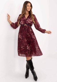 Bordeauxrotes plissiertes Kleid mit Blumenmuster, V-Ausschnitt, langen Ärmeln und schwarzem Gürtel, kombiniert mit schwarzen Stiefeln mit hohem Absatz.