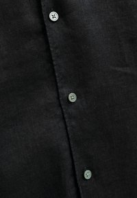 Tissu noir avec trois boutons gris visibles cousus sur une ouverture verticale, montrant un matériau tissé texturé.