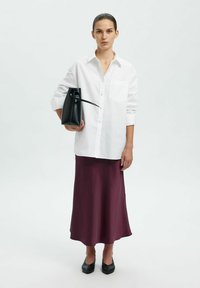 Camicia bianca con bottoni e taschino, abbinata a una gonna di raso bordeaux e scarpe nere. Modella che tiene una borsa nera rigida.
