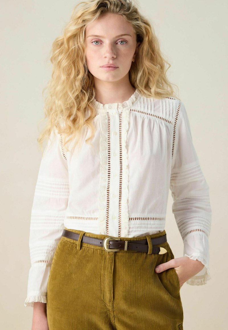 Blouse blanche en tissu texturé, avec des finitions en dentelle et une fermeture à boutons. Associée à un pantalon en velours côtelé vert olive et une ceinture marron.
