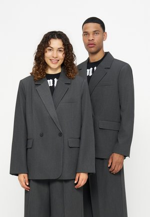 Zwei Models tragen übergroße graue Anzugjacken und passende weite Hosen, die vor einem schlichten weißen Hintergrund stehen.