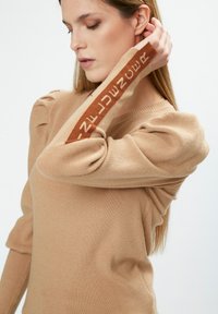 Beige Pullover mit langen Ärmeln, gerippter Textur und gesch pufften Schultern, mit einem kontrastierenden braunen Band mit Text entlang des Arms.