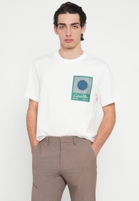 White cotton t-shirt featuring a graphic pocket design with a blue circle and green border. Pairing with light brown tailored trousers.