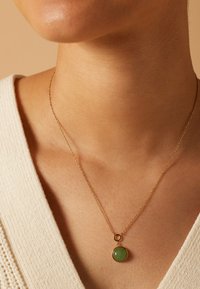 Collier en chaîne en or avec un pendentif en pierre verte ronde, reposant sur un teint de peau, avec un pull crème texturé partiellement visible.