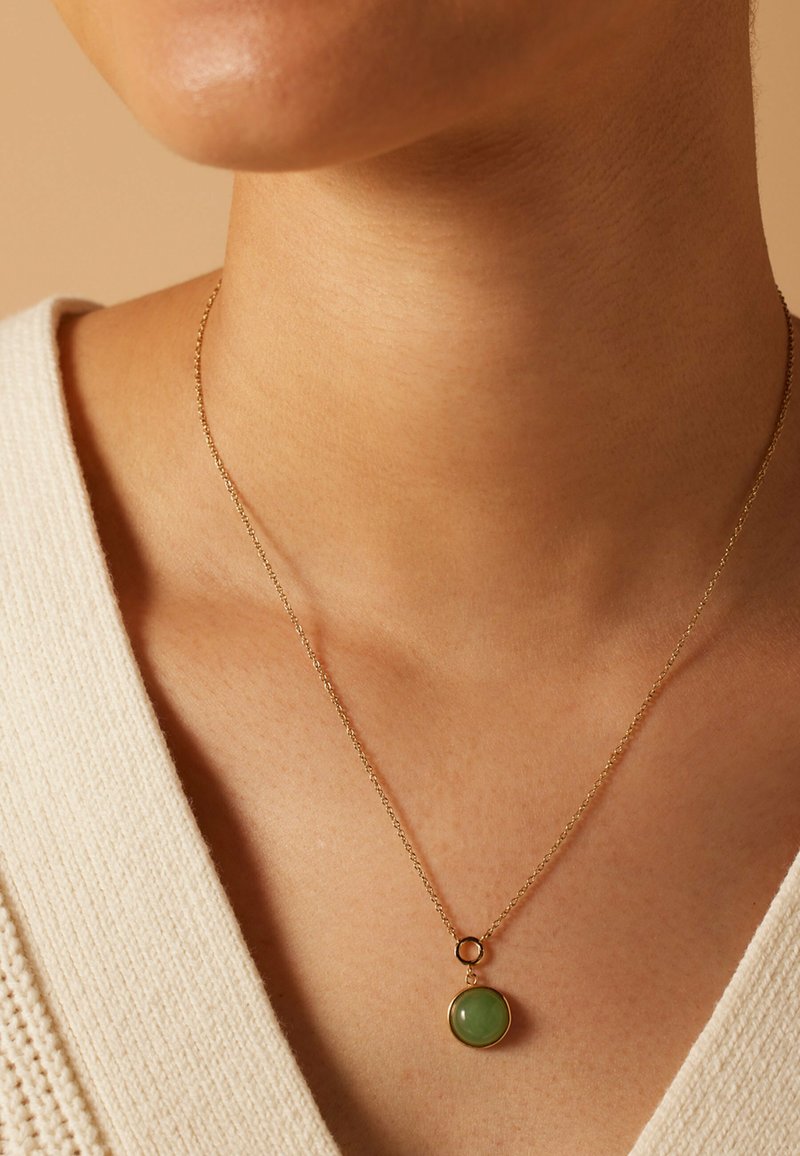 Collier en chaîne en or avec un pendentif en pierre verte ronde, reposant sur un teint de peau, avec un pull crème texturé partiellement visible.