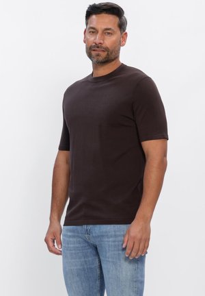 Man met kort donker haar en baard, gekleed in een donkerbruin shirt met korte mouwen en lichtblauwe spijkerbroek, staand tegen een effen witte achtergrond.