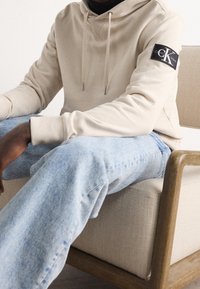 Světlý hnědý svetr s kapucí na tkaničku a logem Calvin Klein na levém rukávu, v kombinaci se světle modrými džínami.