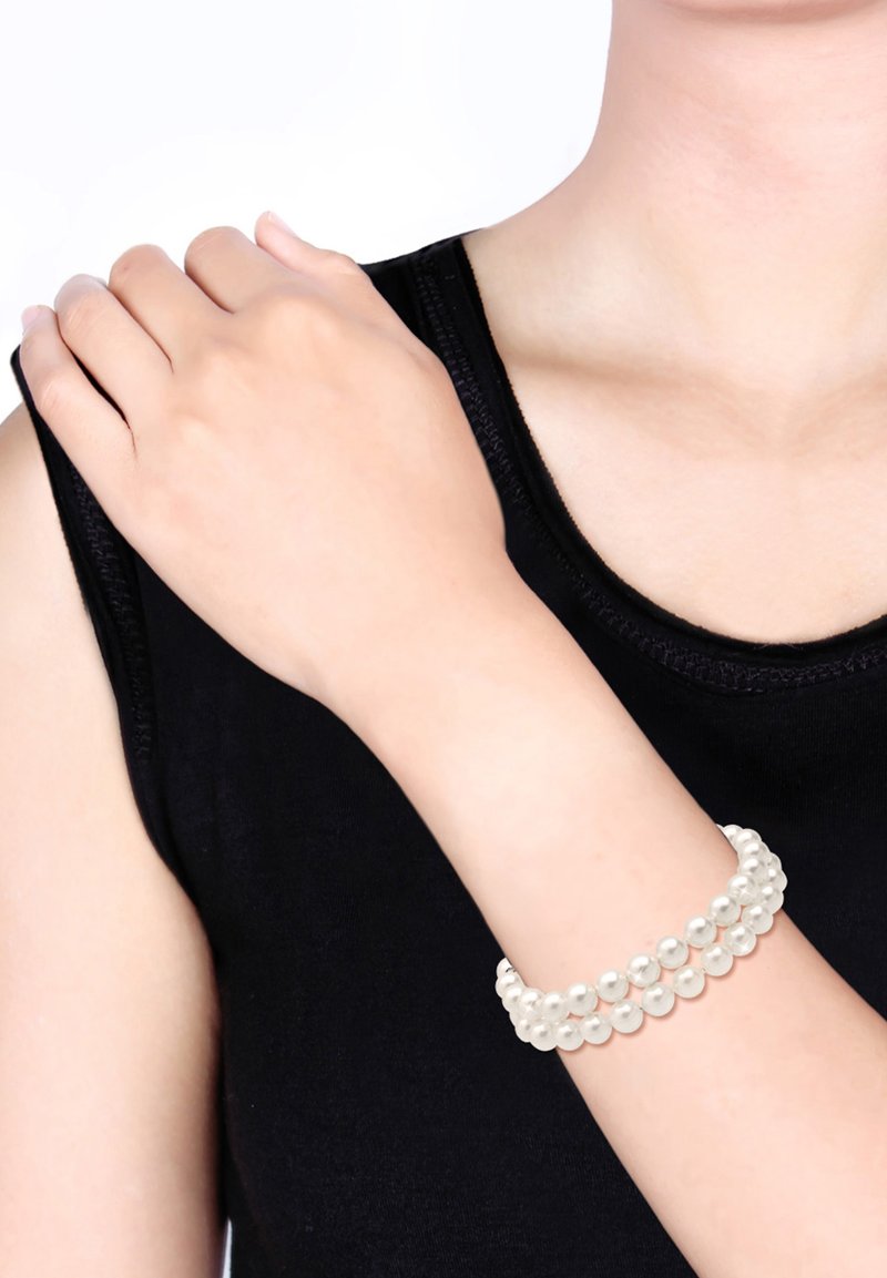 Weißes Perlenarmband mit mehreren Strängen, das auf einem Handgelenk ruht. Der Hintergrund ist ein einheitliches Weiß, und der Arm ist nackt mit einem schwarzen Kleidungsstück.