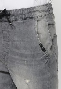 Sive traper shorts s pojasom na vezanje, bočnim džepovima i izraženim istrošenim detaljima. Sadrži crnu logotip oznaku na džepu.