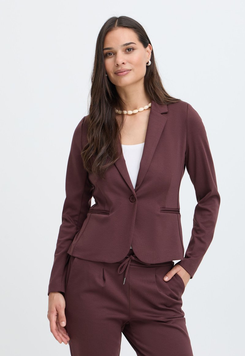 Femme aux longs cheveux foncés portant un collier de perles, un blazer bordeaux et un pantalon assorti, debout avec une main dans la poche devant un fond uni.