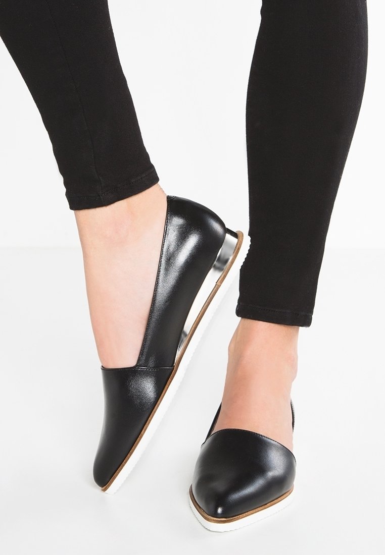 Czarne skórzane mokasyny na płaskim obcasie z ostrym noskiem. Biała śródpodeszwa i gładka tekstura podkreślają design. Noszone z czarnymi spodniami.